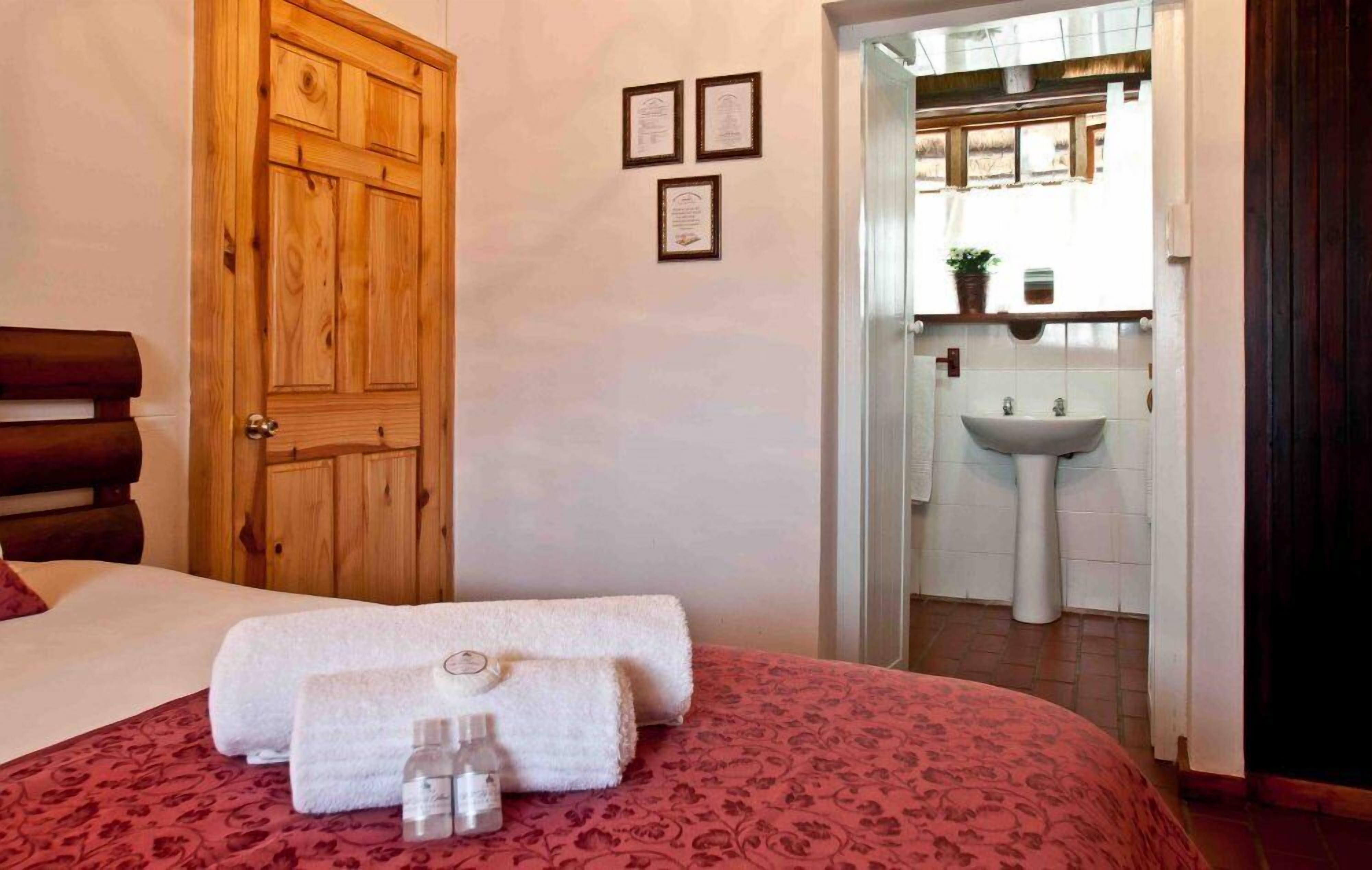 Habitación de hotel con puerta de madera, cama con colcha estampada roja, dos toallas enrolladas y una botella pequeña sobre la cama.