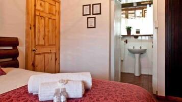 Habitación de hotel con puerta de madera, cama con colcha estampada roja, dos toallas enrolladas y una botella pequeña sobre la cama.