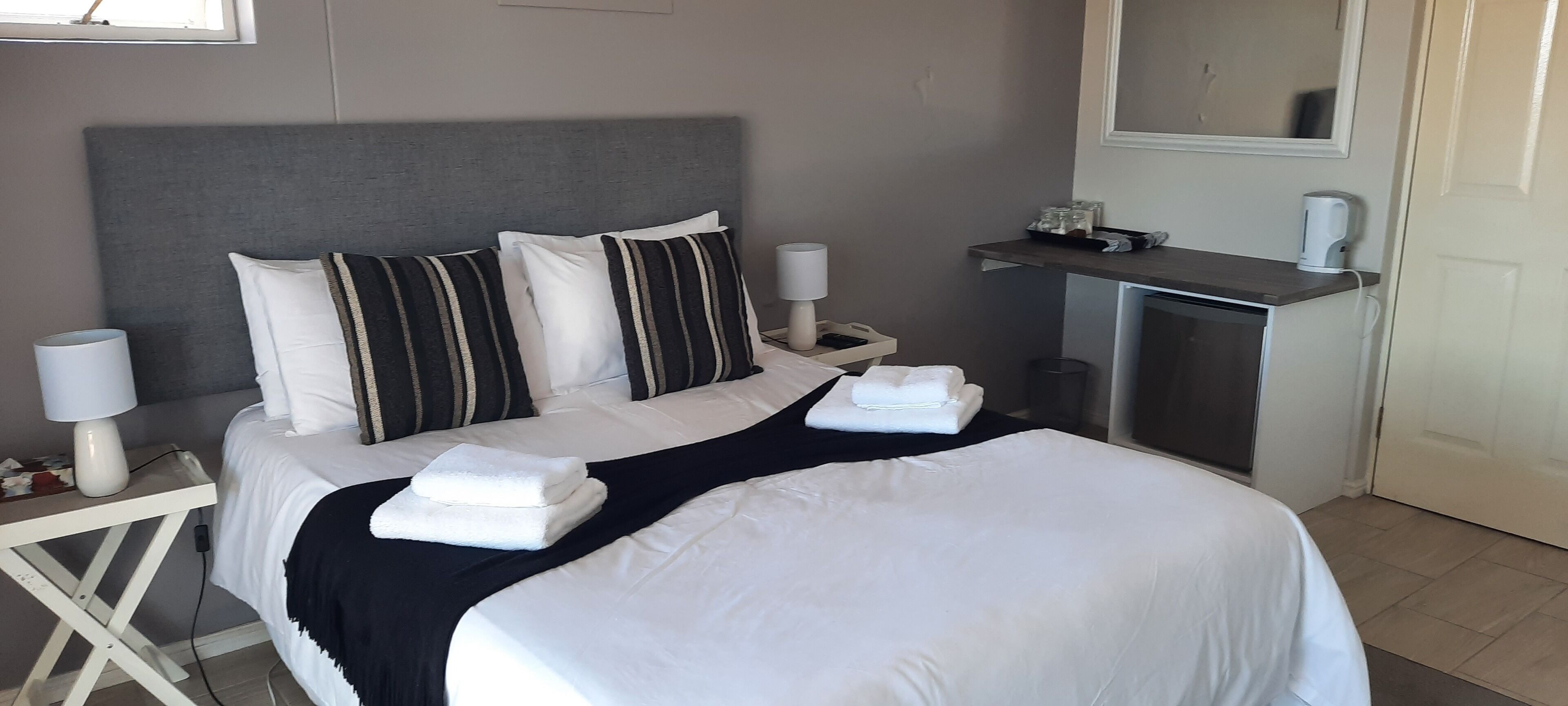 A neatly made bed with white linens and striped pillows, a bedside table with a lamp, and a small desk with a mirror and a kettle.