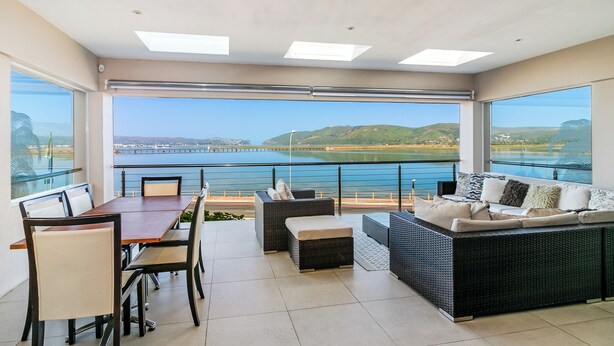 A balcony with a dining table and chairs, a sofa, and a view of the sea and hills. - Phoenix Lodge and Waterside Accommodation (Knysna)