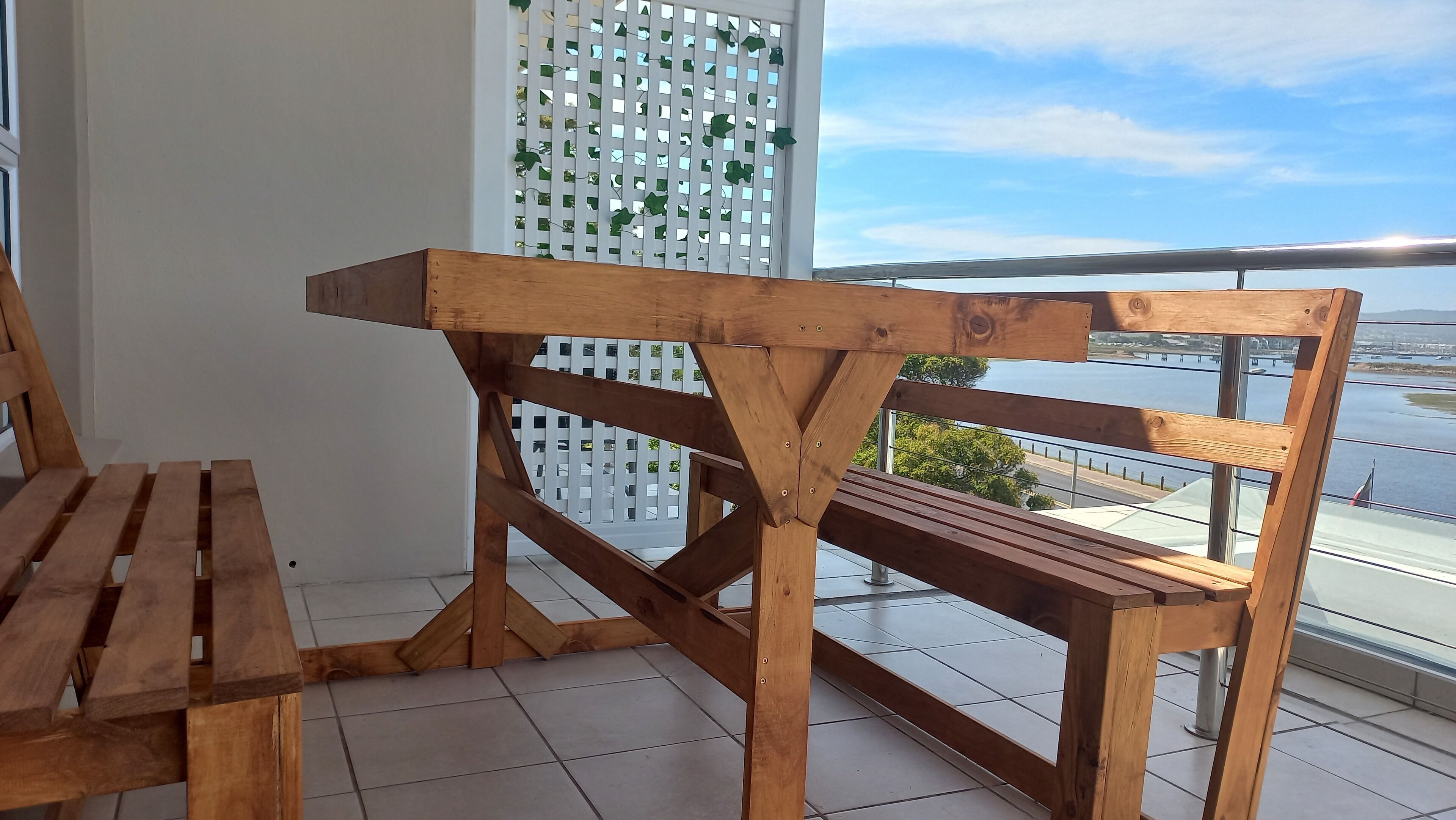 A wooden bench with a backrest and a table with a wooden top, positioned on a balcony with a view of a body of water and a white fence with green foliage.