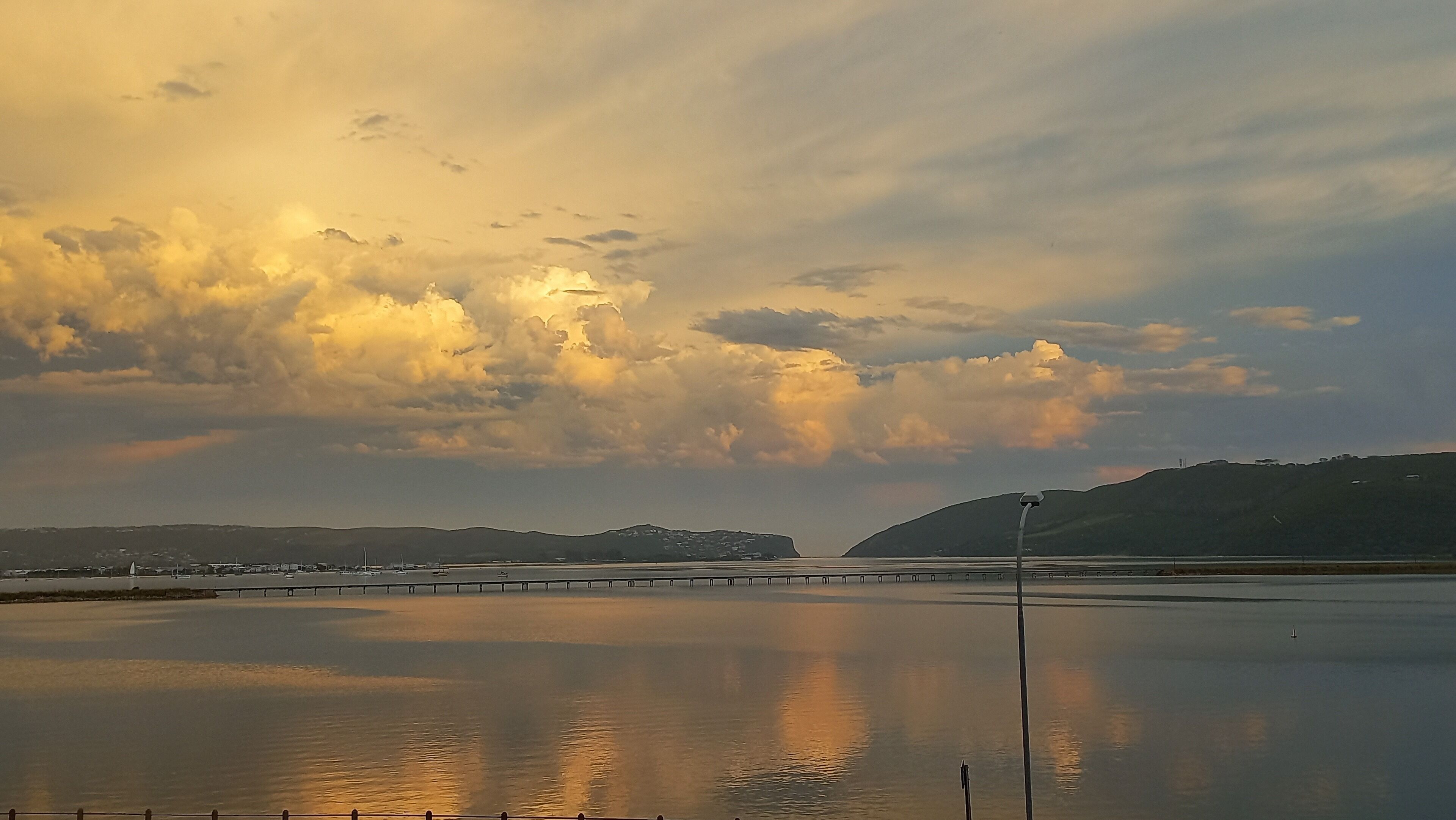 A calm body of water reflecting clouds and a bridge in the distance.