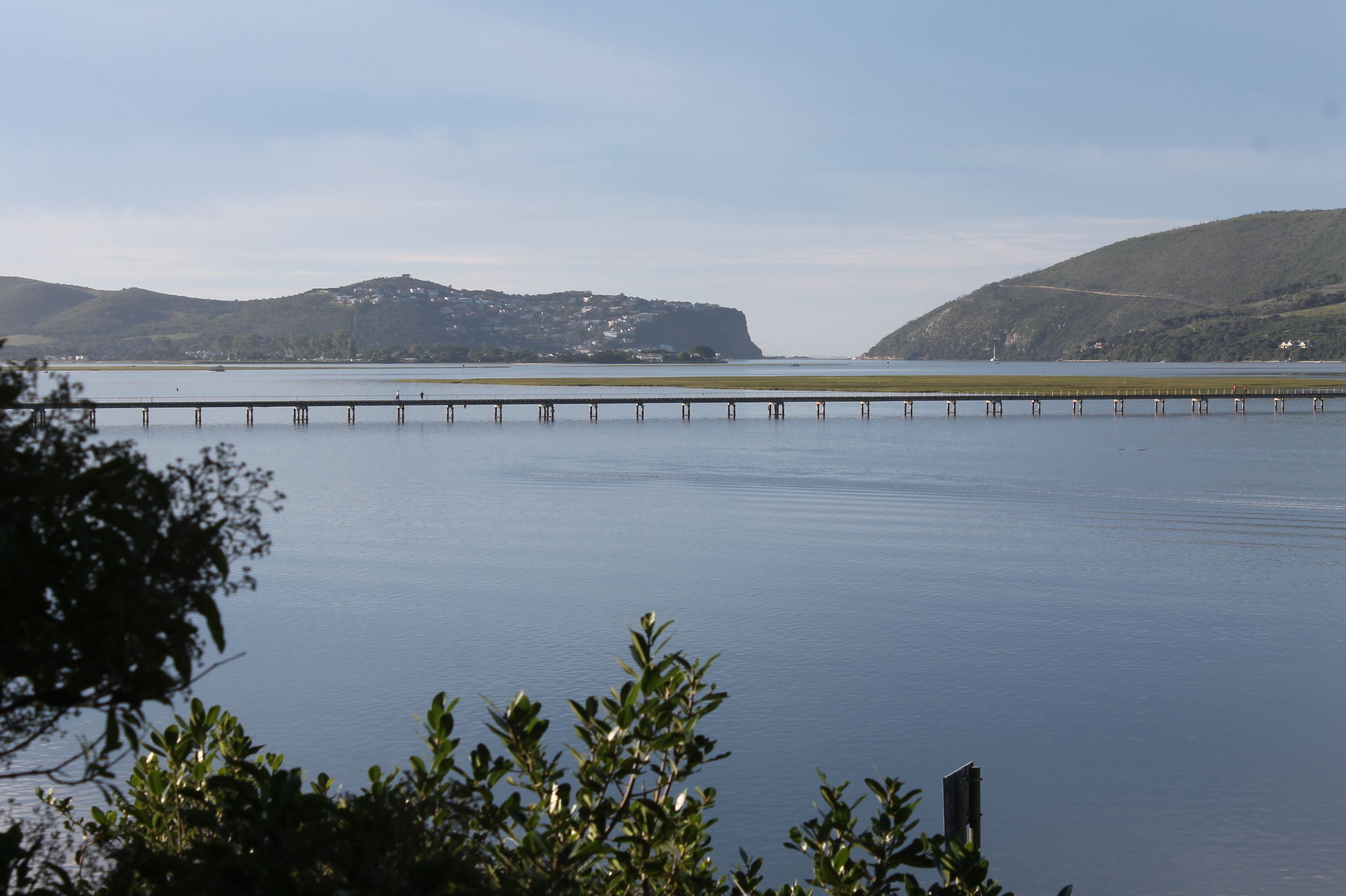 A calm lake with a pier extending into the water, surrounded by hills and sparse vegetation.