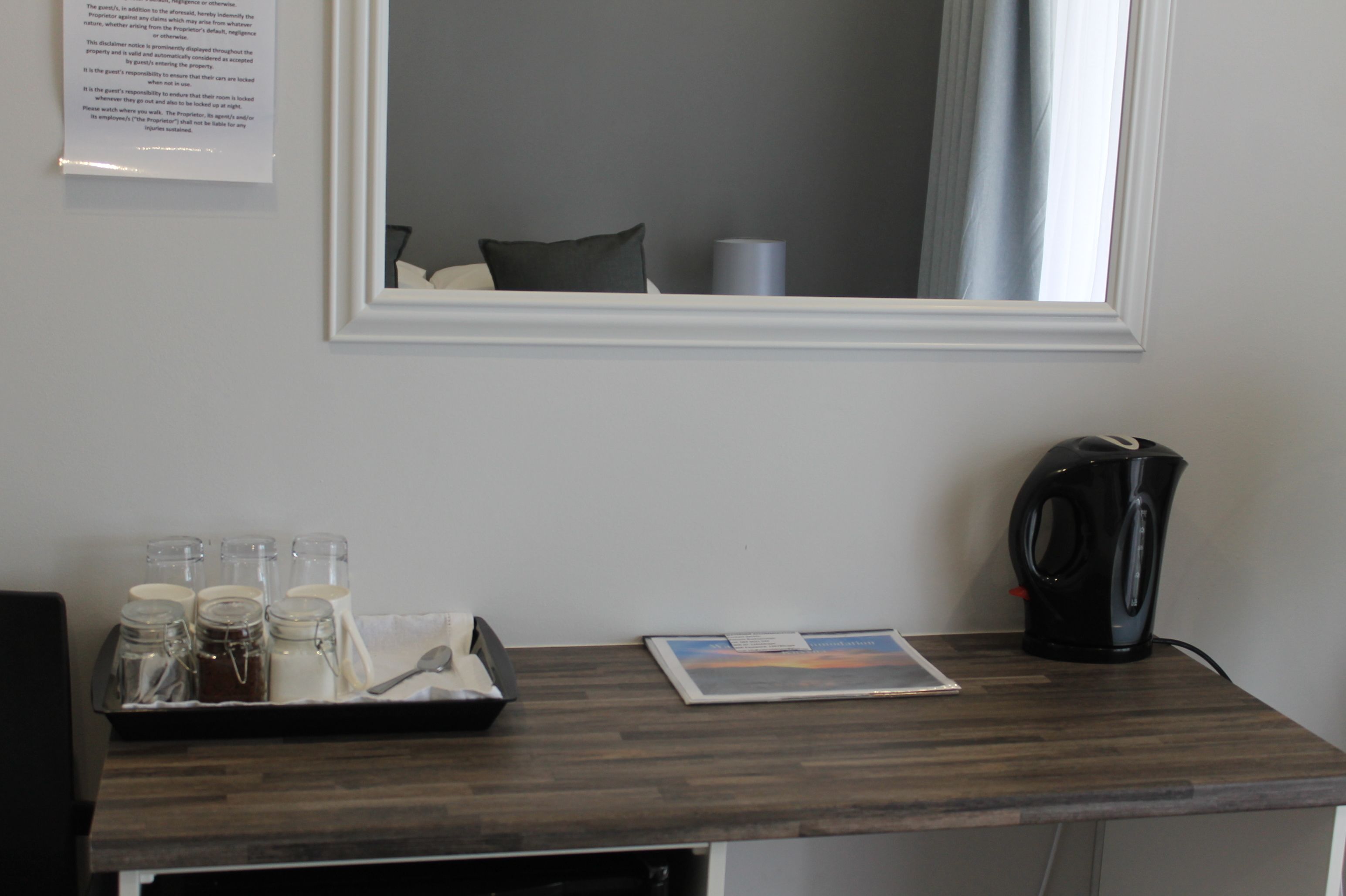 A hotel room with a wooden desk, a kettle, a tray with glass jars, and a framed picture.