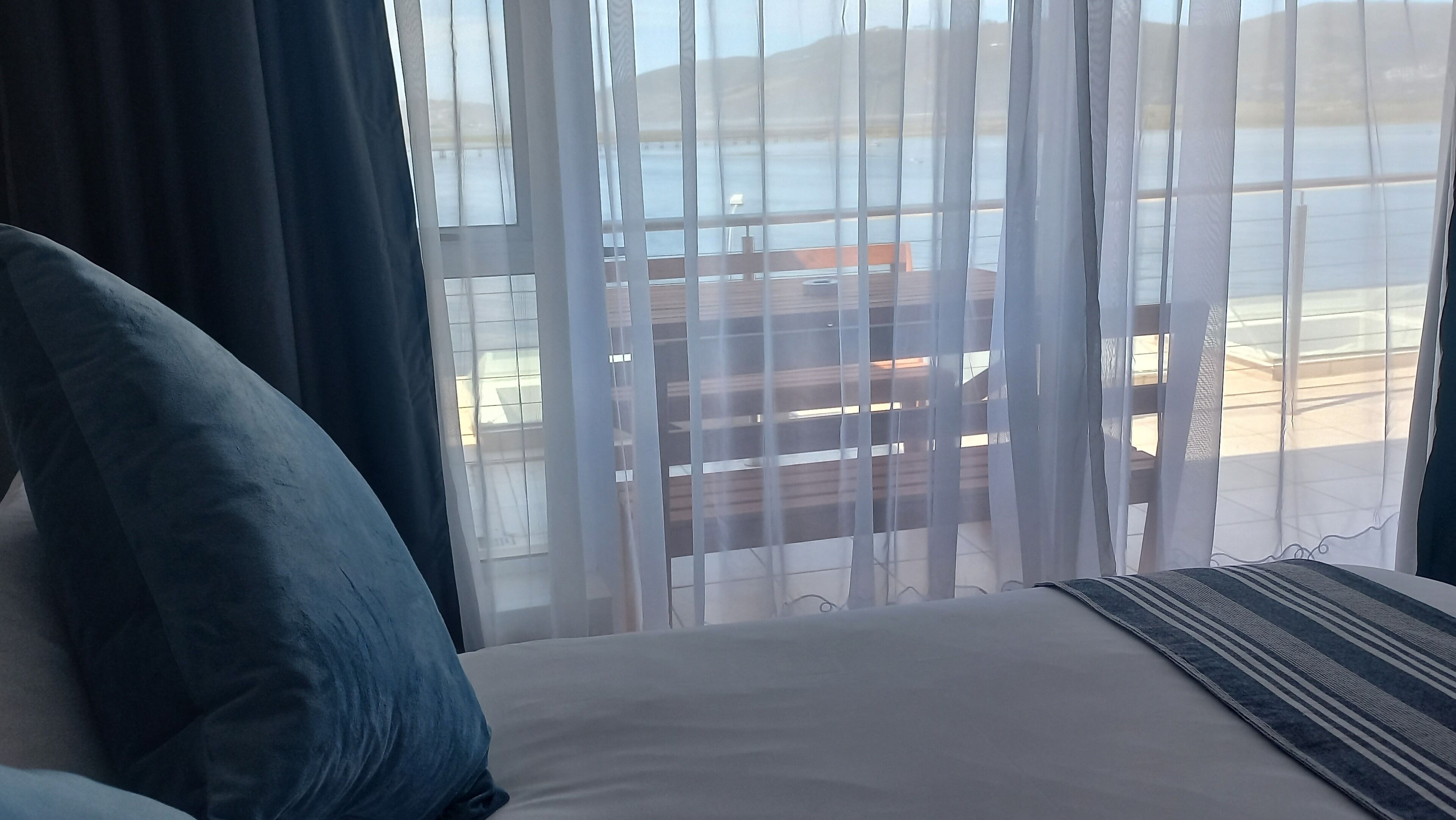 Habitación de hotel con cama, vista al mar a través de cortinas translúcidas y una terraza de madera afuera.