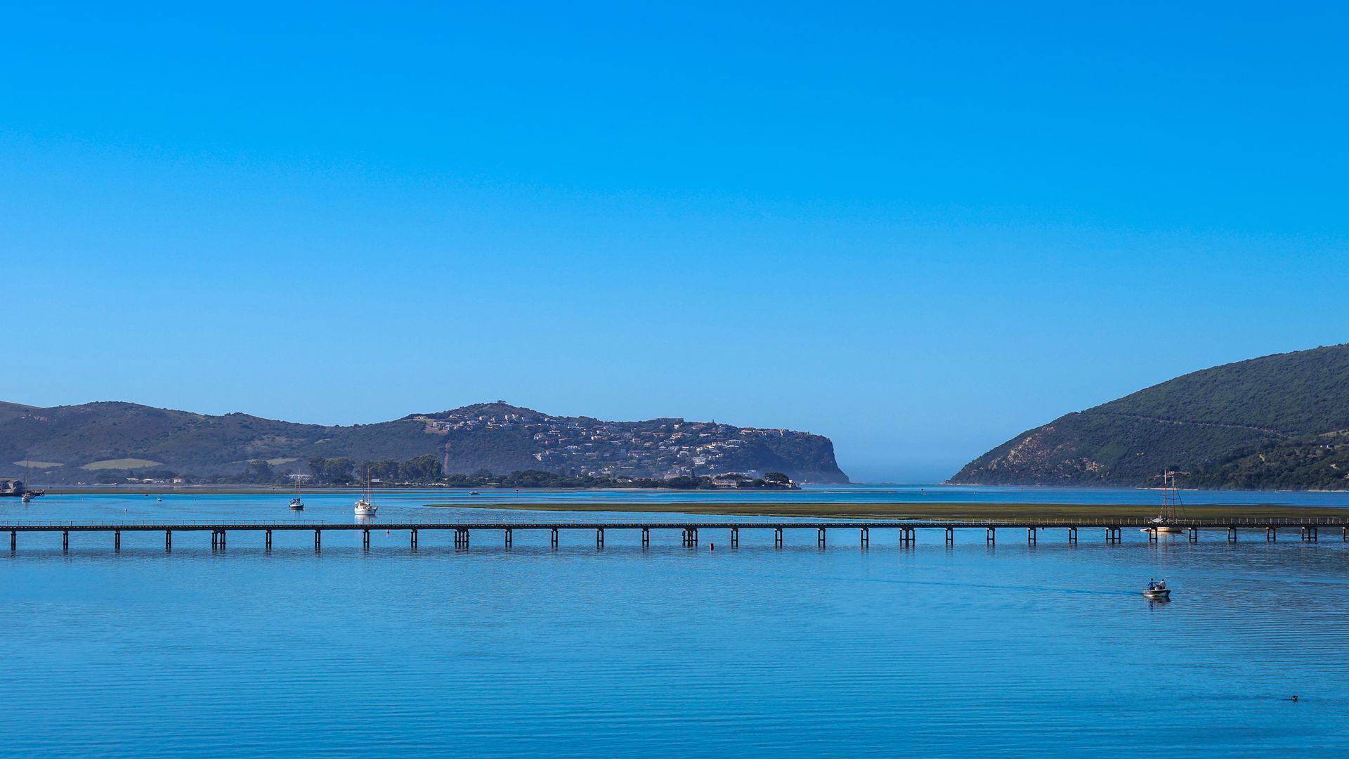 A coastal scene with a pier extending into calm blue waters, surrounded by hills and a clear sky.