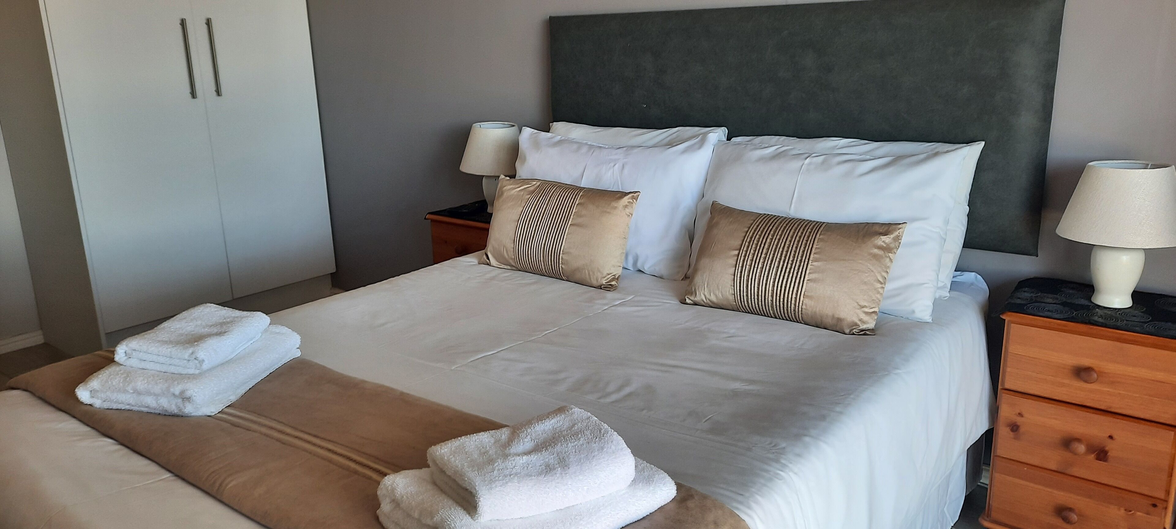 A neatly made bed with white linens and two beige pillows, a wooden nightstand with a lamp, and a wardrobe with two doors.