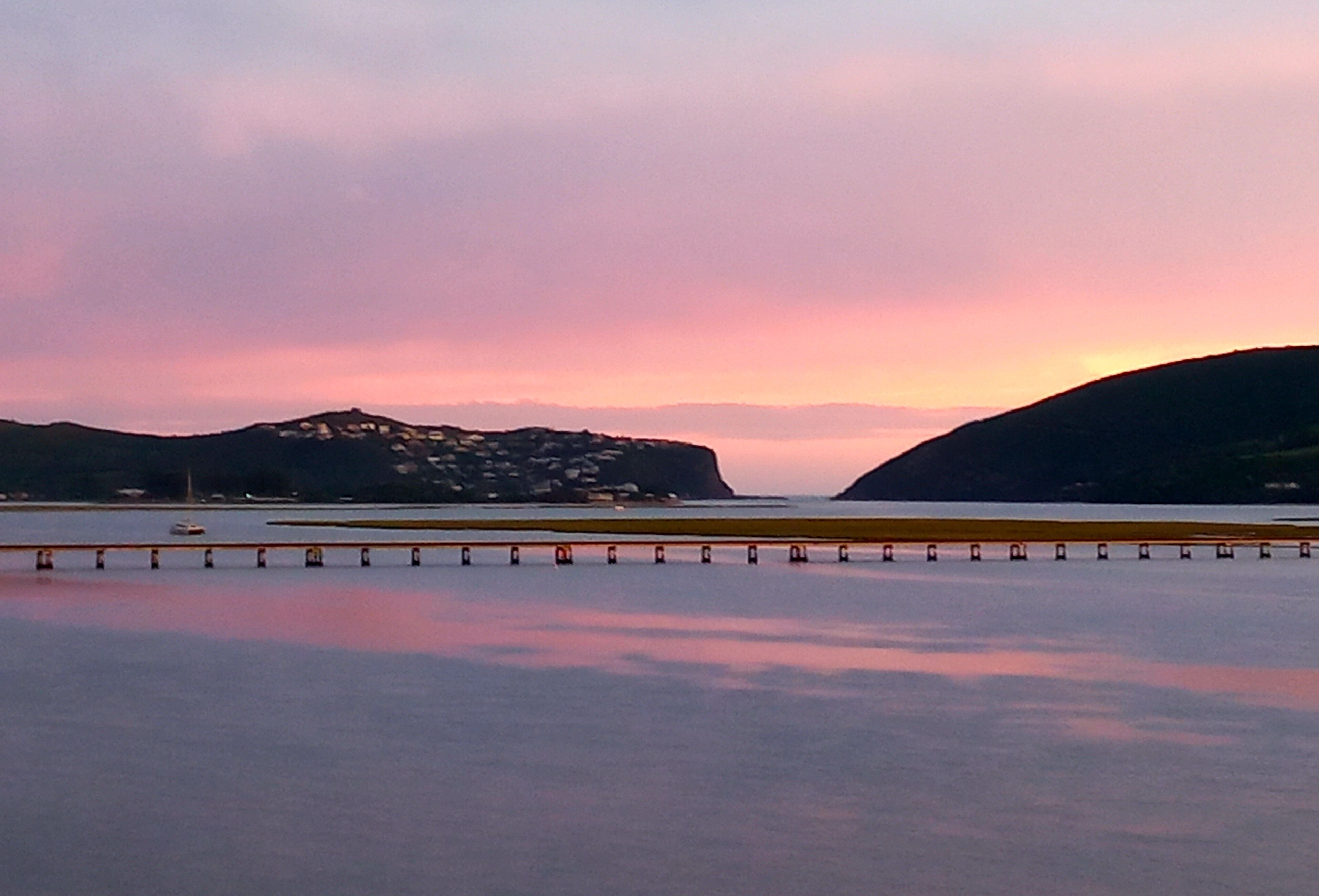 A coastal landscape with a calm sea, a bridge, and distant hills.