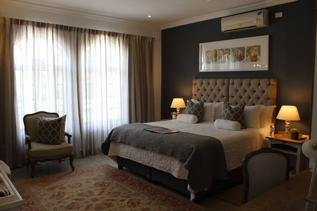 Ein Hotelzimmer mit einem großen Bett, zwei Nachttischen mit Lampen, einem Sessel, einem Schreibtisch und einem großen Fenster mit Vorhängen.