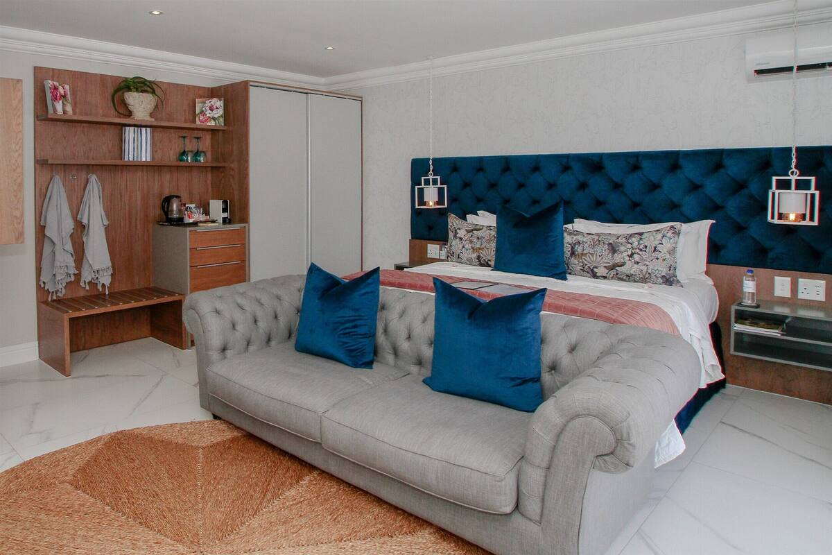 Ein modernes Schlafzimmer mit grauem Sofa, einem großen Bett mit blau-blumigen Kissen, einem Holzschrank und einer Pendelleuchte.