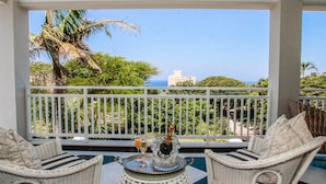 A balcony with wicker furniture, a table with a bottle and glasses, and a view of the ocean and greenery.