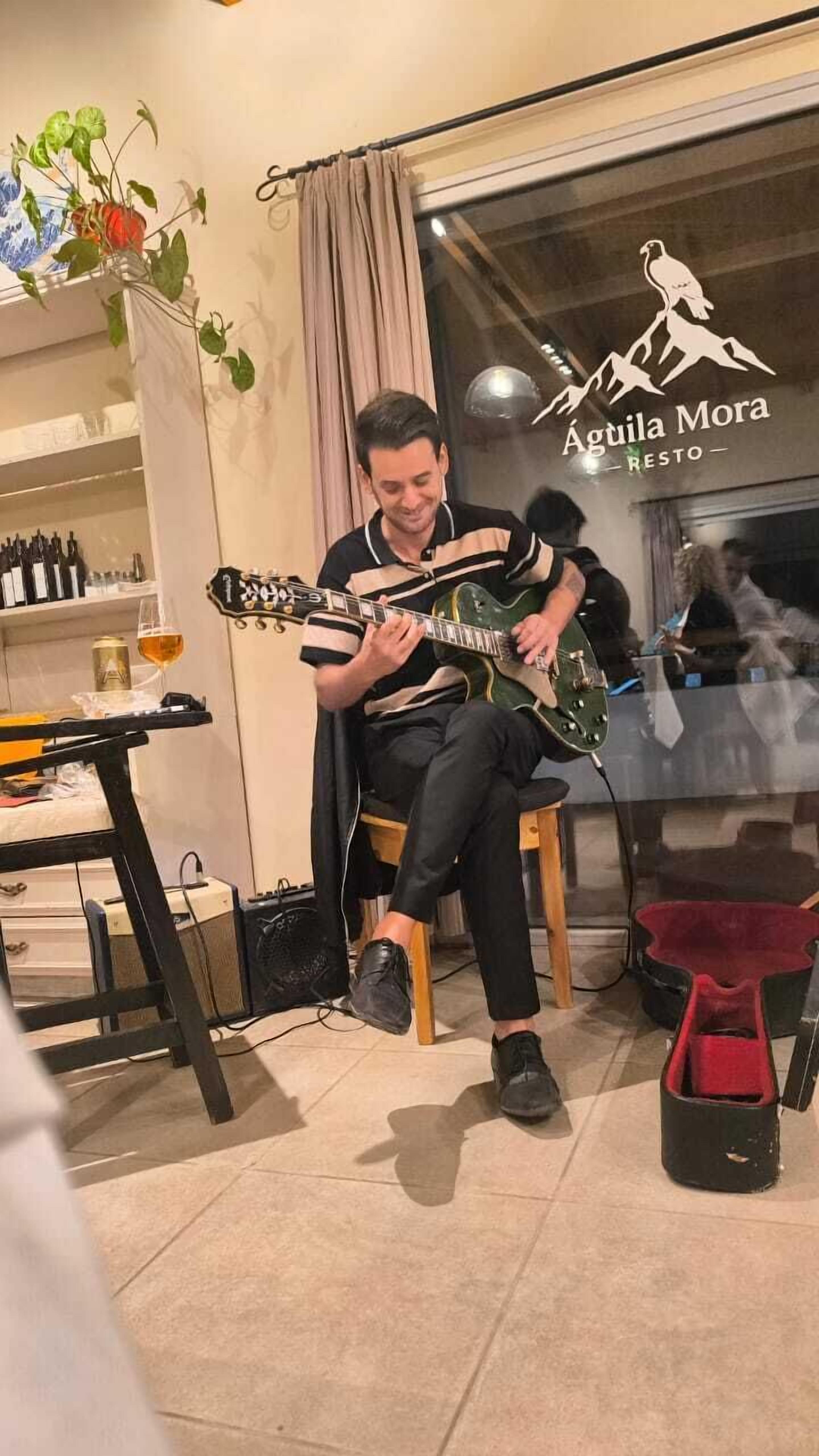 Eine Person spielt Gitarre in einem Raum mit Tisch, Weinflasche und Pflanze.