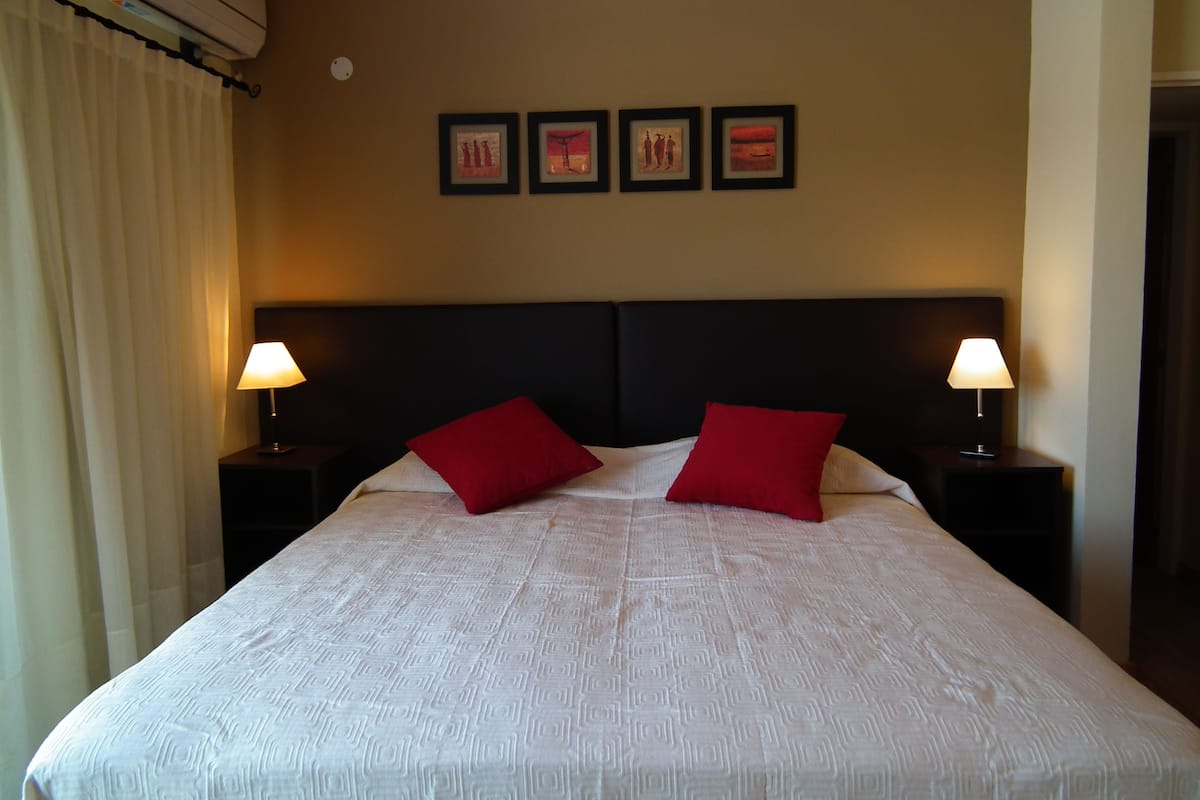 Een hotelkamer met een groot bed, twee rode kussens, een donker hoofdeinde en twee bedlampjes.