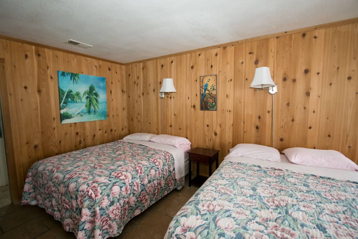 Quarto com duas camas, paredes de painel de madeira, um quadro de uma praia e duas luminárias fixadas na parede.