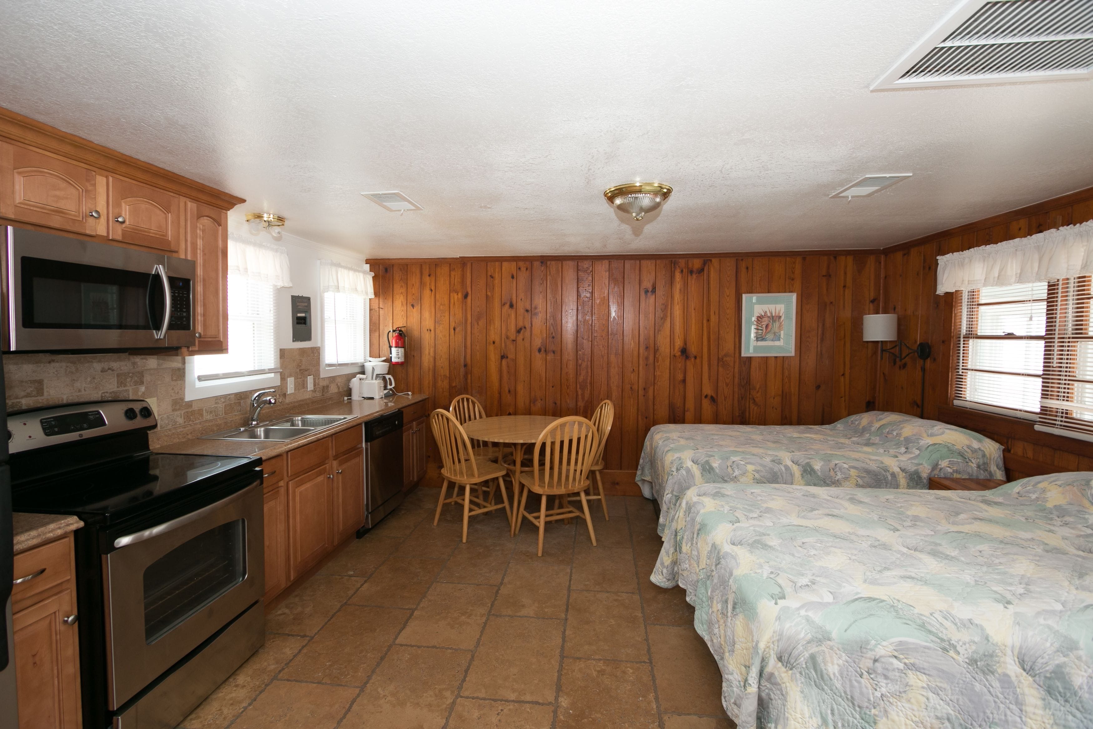 A compact kitchen with a microwave, stove, and sink, wooden cabinets, and a dining table with chairs. There are two beds with floral bedding.
