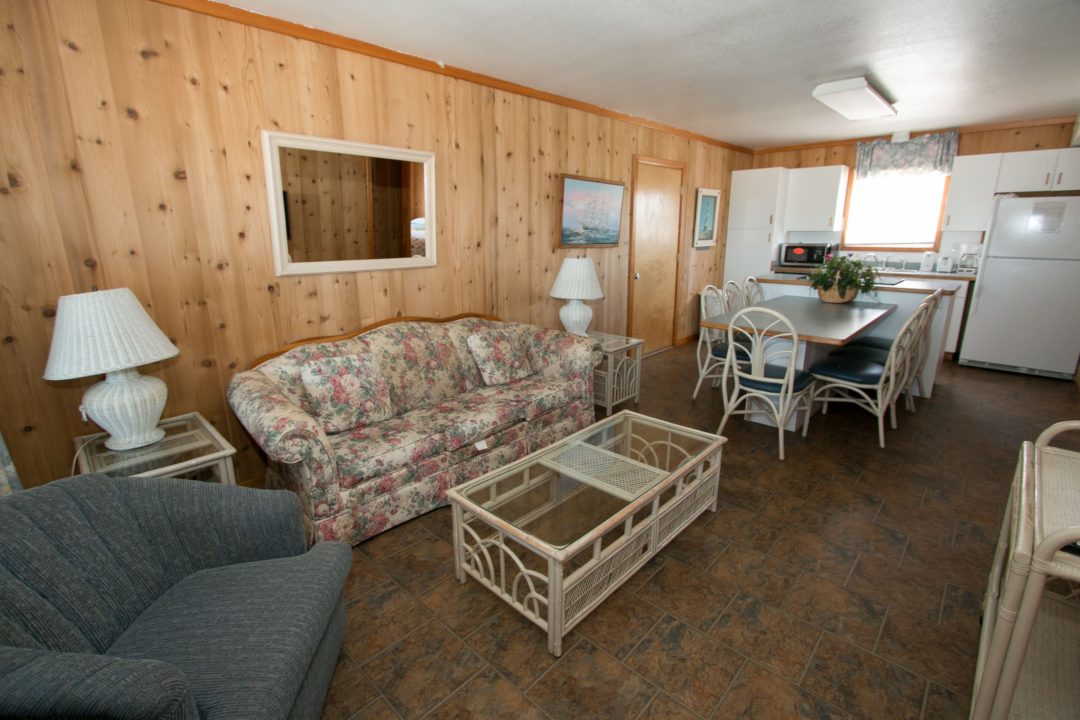A compact living space with a sofa, a coffee table, a dining table with chairs, a kitchen area with a refrigerator, and a window with curtains.