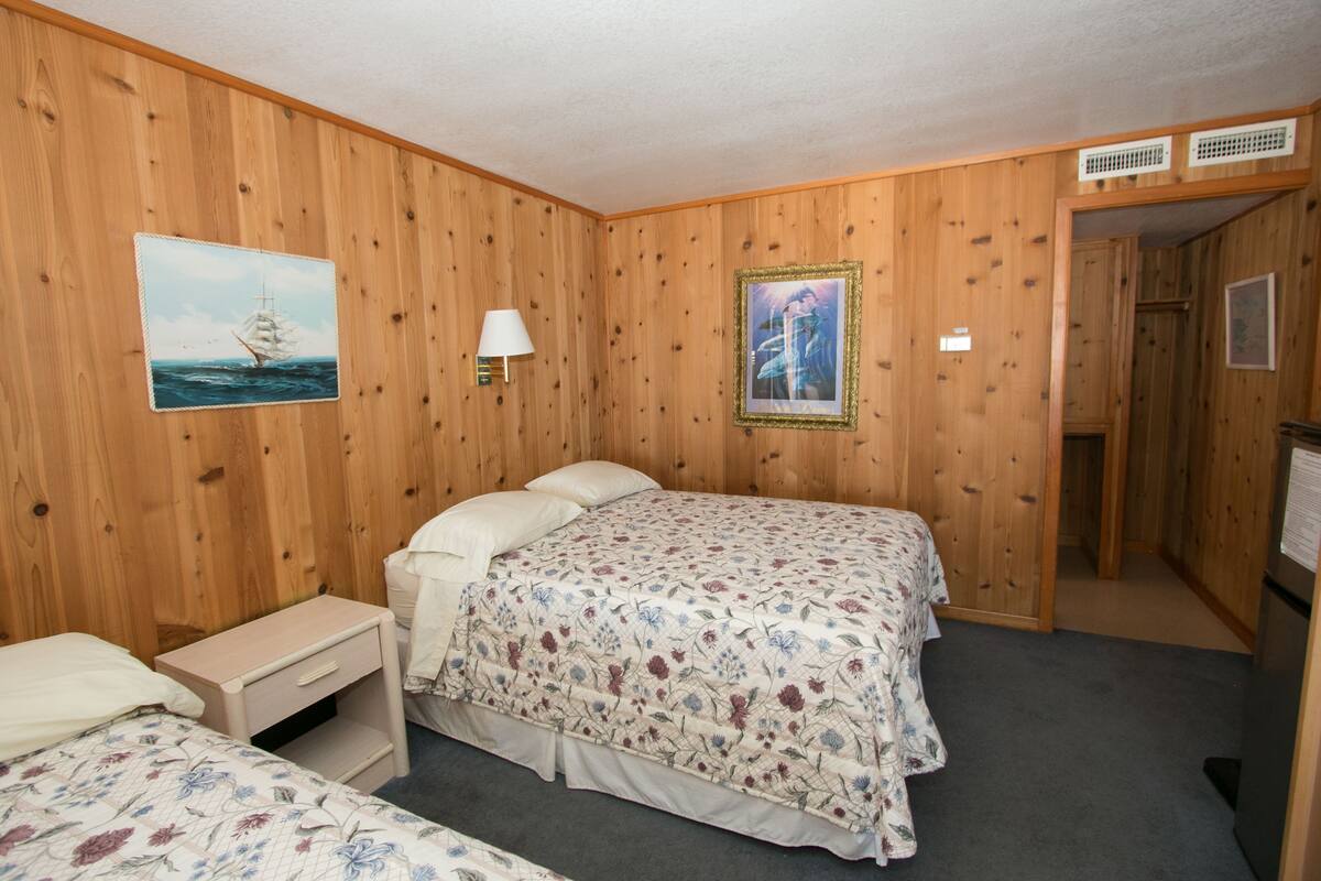 Quarto com painéis de madeira, uma cama, uma mesa de cabeceira, um abajur e um quadro na parede.