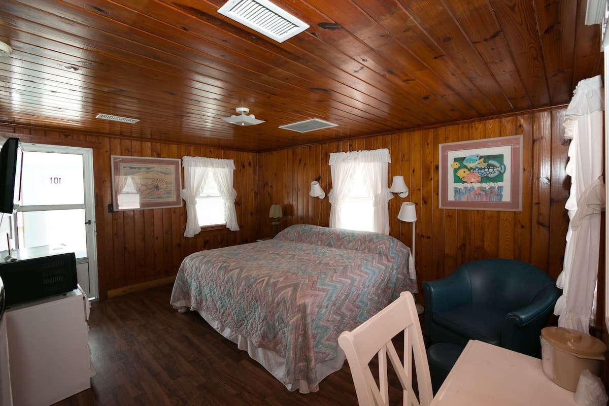 Quarto com painéis de madeira, uma cama, uma escrivaninha, uma cadeira e uma janela com cortinas.