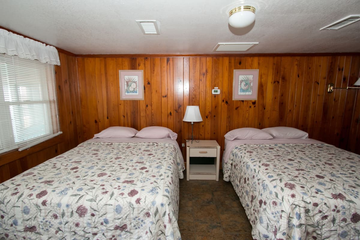 Quarto com paredes de madeira, duas camas com roupa de cama florida, mesa de cabeceira, luminária fixada na parede e termostato.