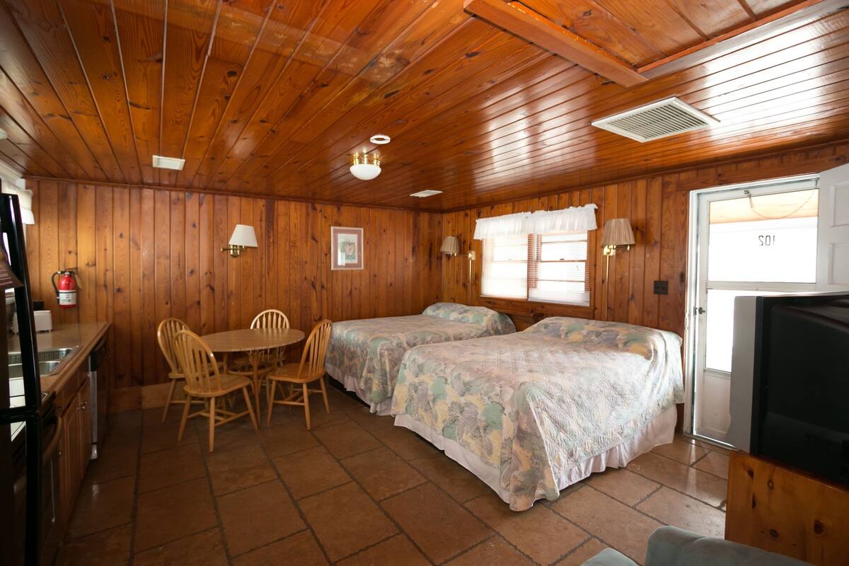 Uma cabana de madeira com duas camas, uma mesa de jantar, cadeiras, uma televisão e uma porta com a inscrição 'SOI'.