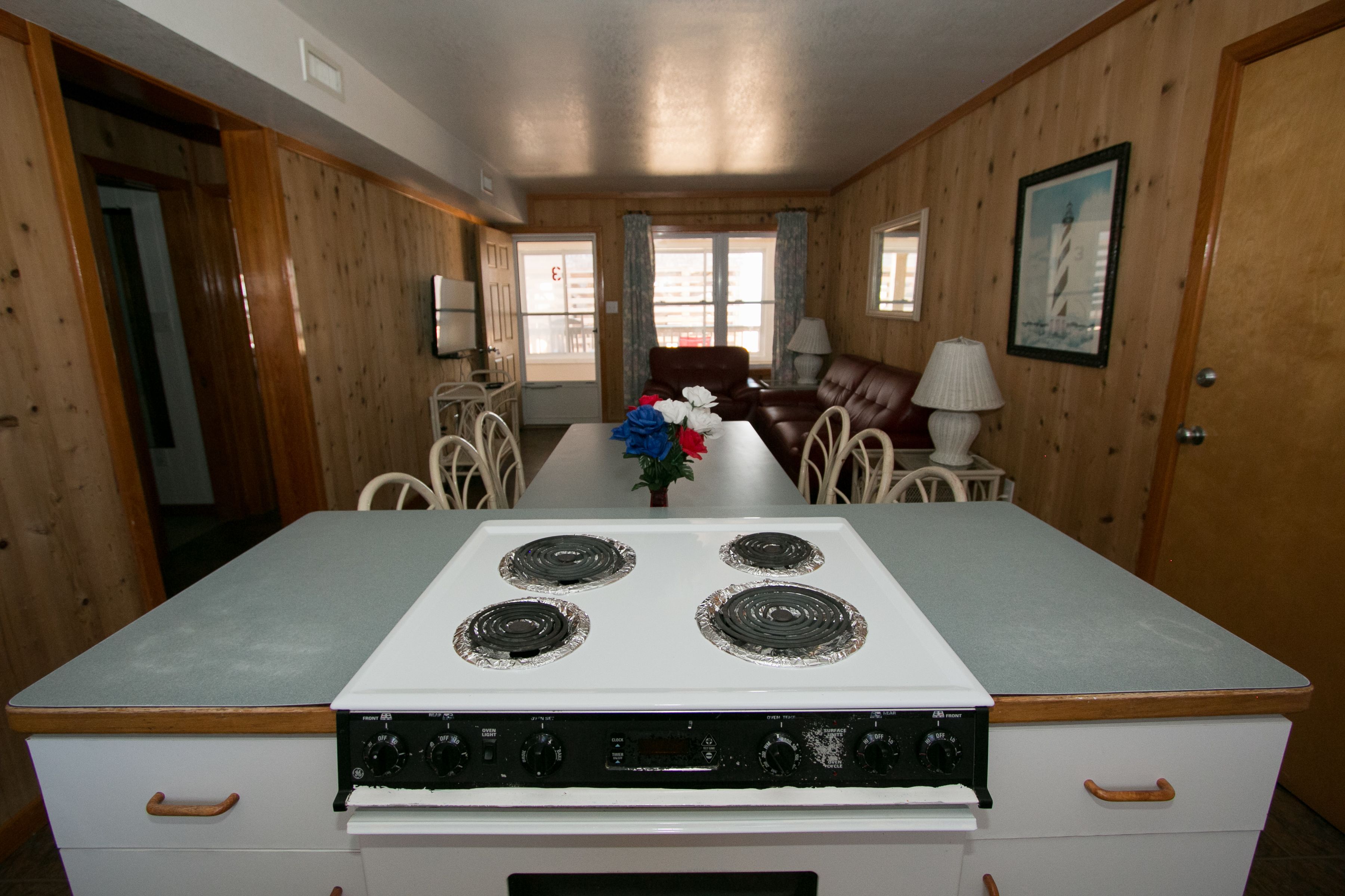 A kitchen with a stove, dining table, chairs, and a view of the outside through large windows.