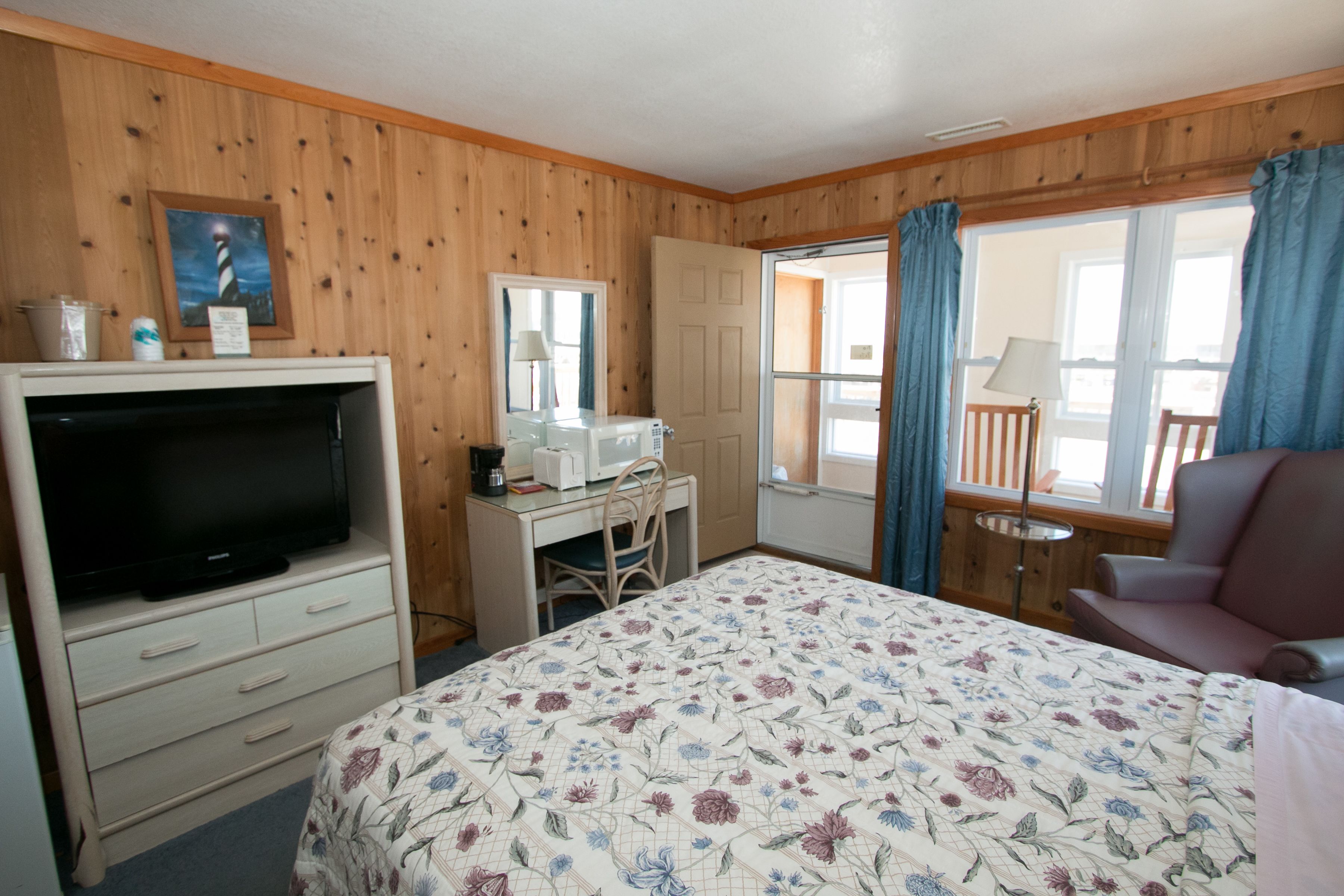 Quarto com cama, televisão, escrivaninha, cadeira e janela com cortinas.