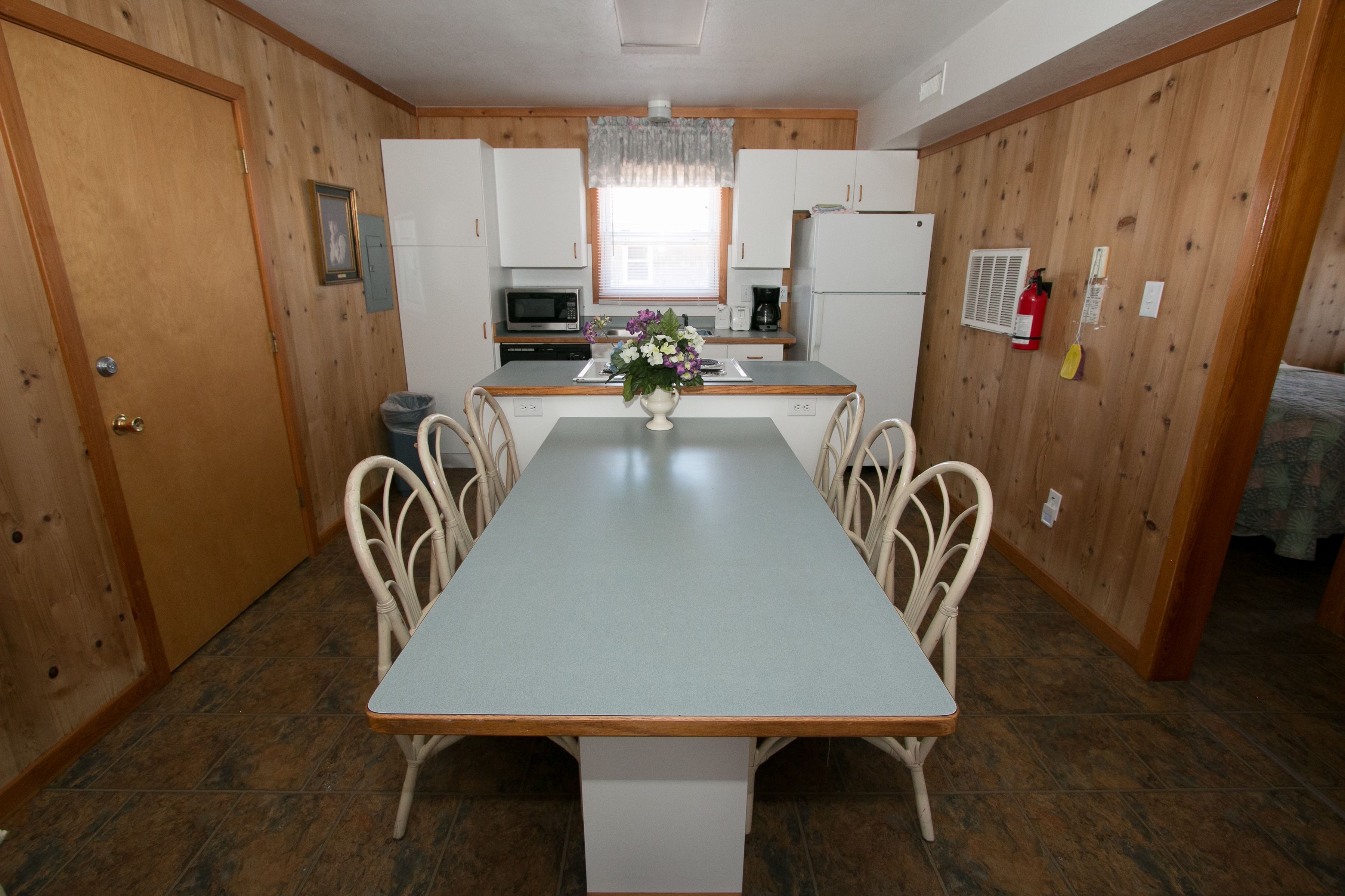 Una cocina funcional con mesa, sillas blancas, microondas, refrigerador y ventana con cortina.