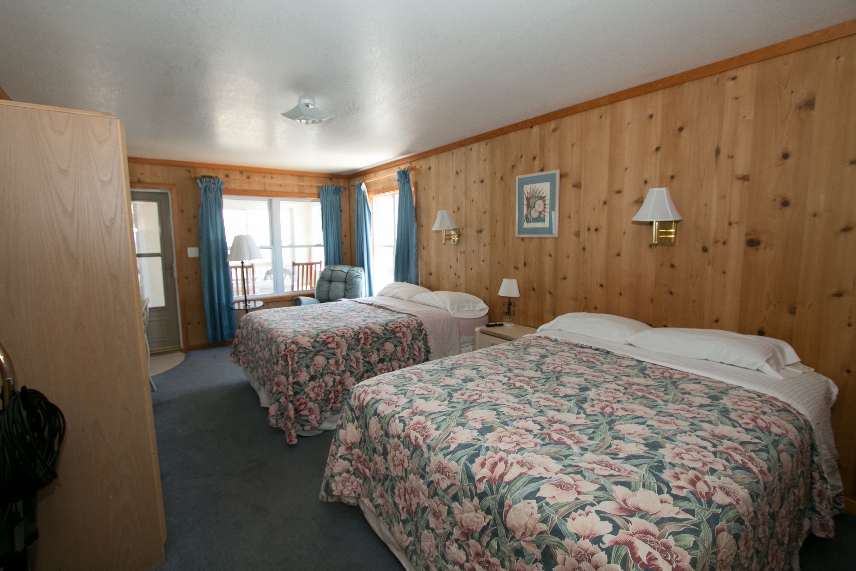 A room with two beds, wooden walls, a window with curtains, a chair, and a table.
