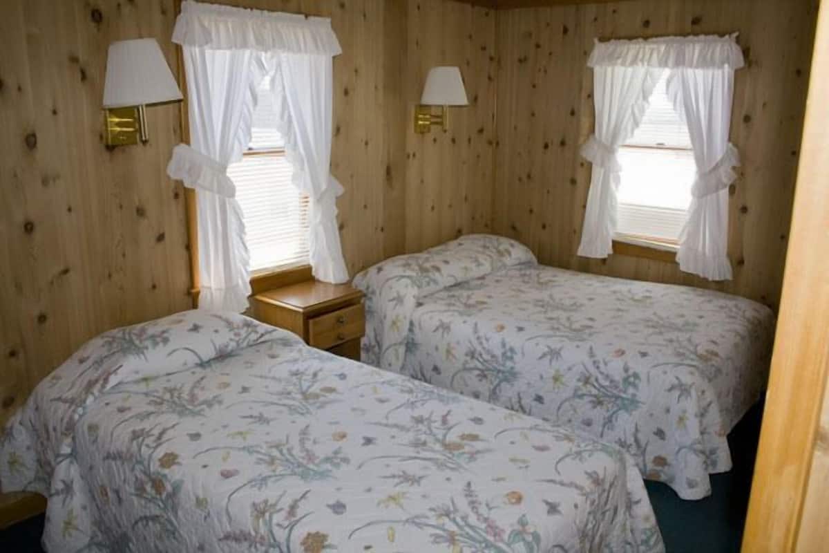 Quarto com duas camas, cada uma com colcha florida e cortinas brancas. Há duas luminárias fixadas na parede e uma pequena mesa de cabeceira de madeira entre as camas.