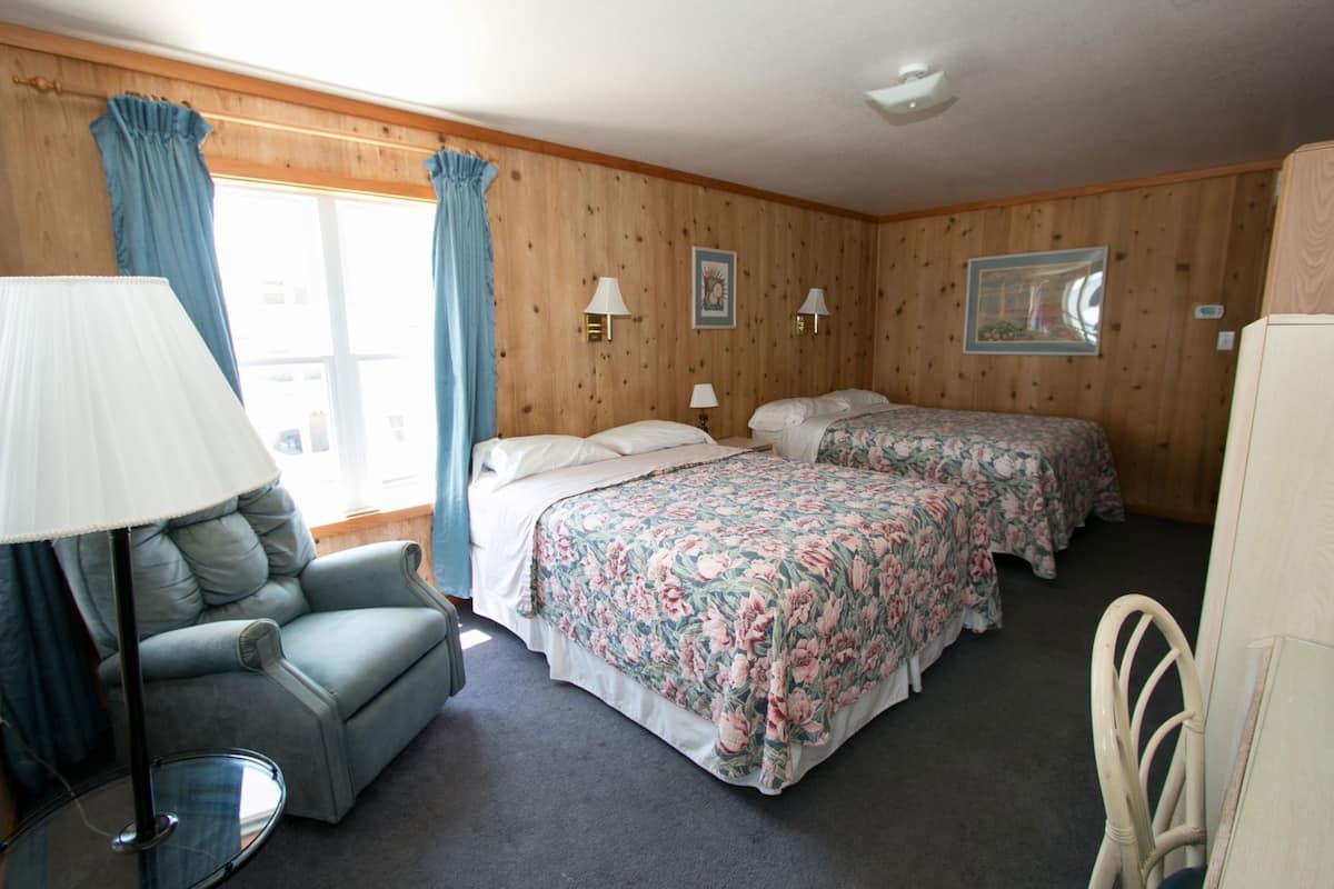 Quarto com duas camas, uma cadeira, um abajur e uma janela com cortinas.
