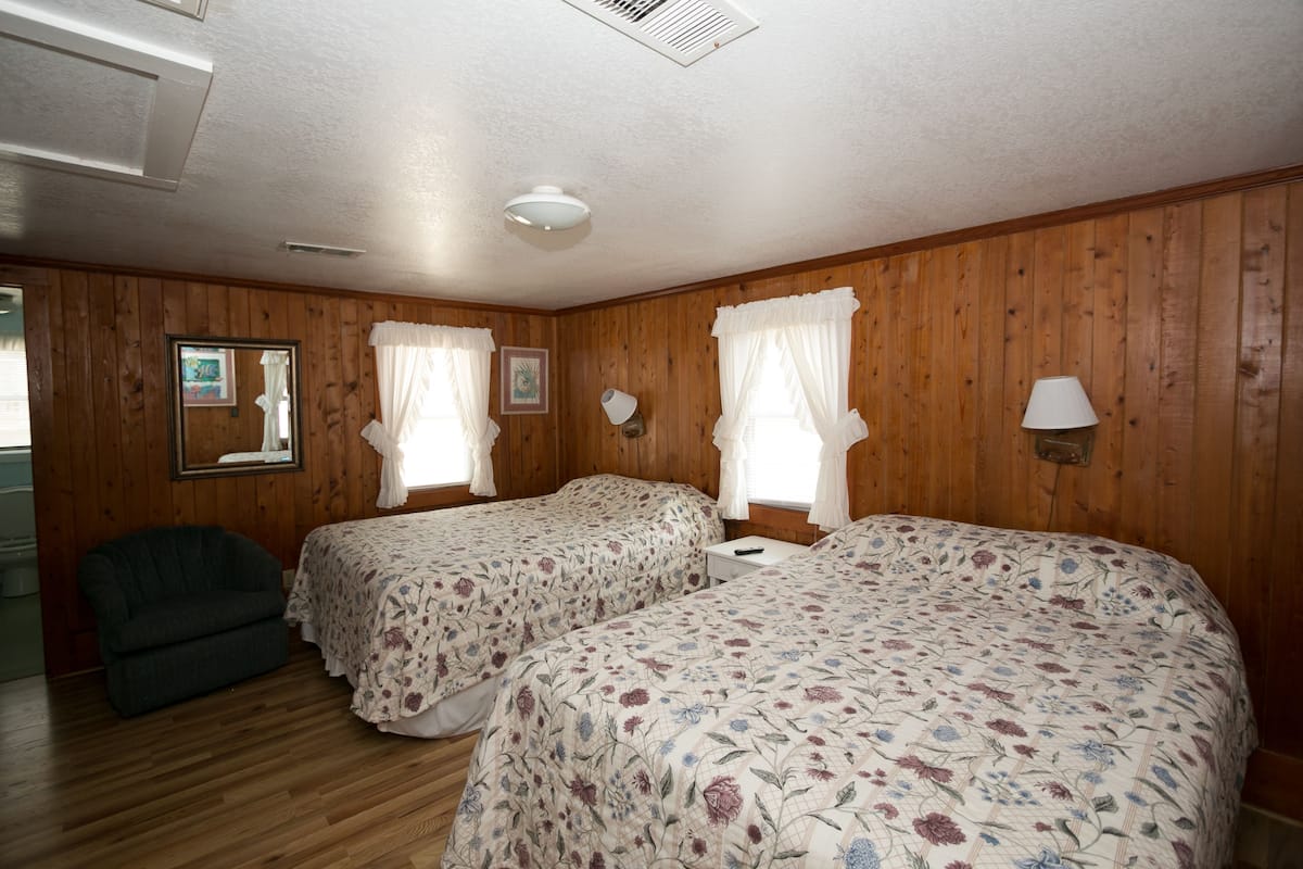 Quarto com painéis de madeira, duas camas com roupa de cama floral, uma mesa de cabeceira, um abajur e uma cadeira.