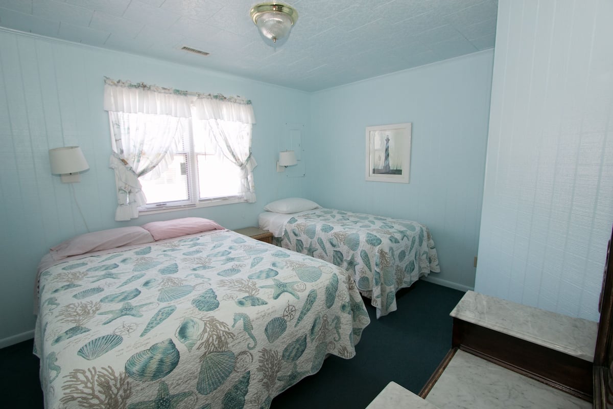 Quarto com duas camas, janela com cortinas, luminária na parede e um quadro emoldurado.