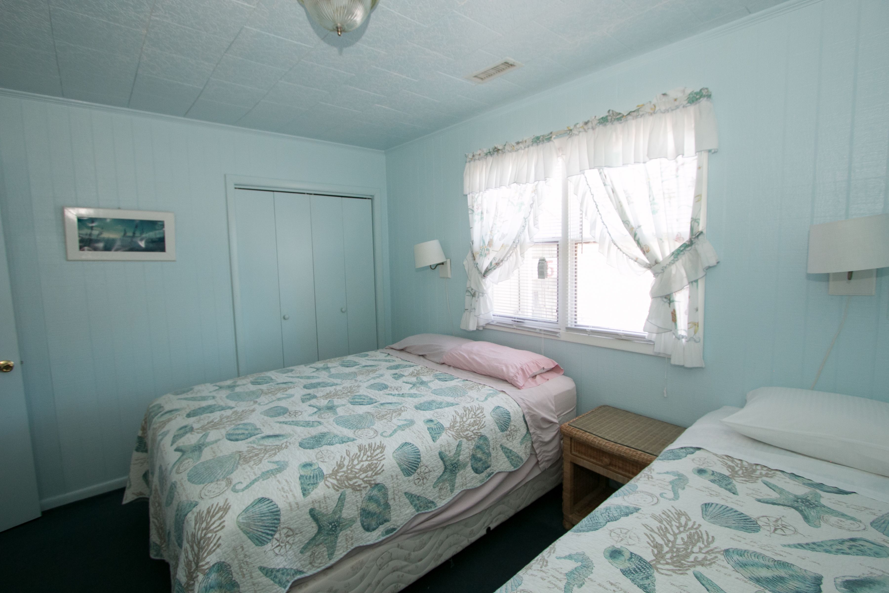 A bedroom with two beds, a window with curtains, a bedside lamp, and a framed picture on the wall.