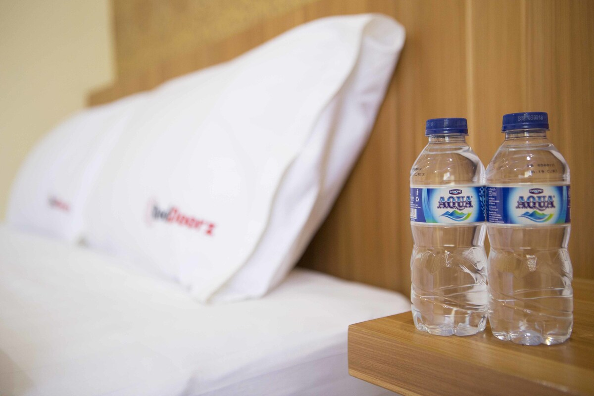 Sängyssä on valkoiset vuodevaatteet ja yöpöydällä, joka on tehty puusta, on kaksi Aquafina-vesipulloa.