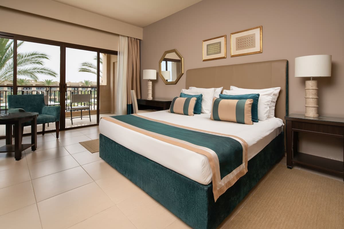 Ein Hotelzimmer mit einem großen Bett, einem Schreibtisch, einem Stuhl, einem Balkon mit Blick auf Palmen und zwei gerahmten Bildern an der Wand.