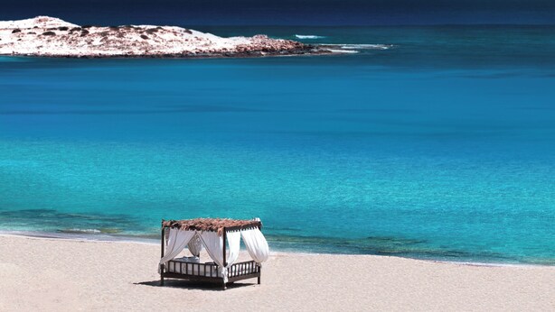 Una cama individual con dosel en una playa arenosa, con vista a un mar azul cristalino y un promontorio rocoso a lo lejos.