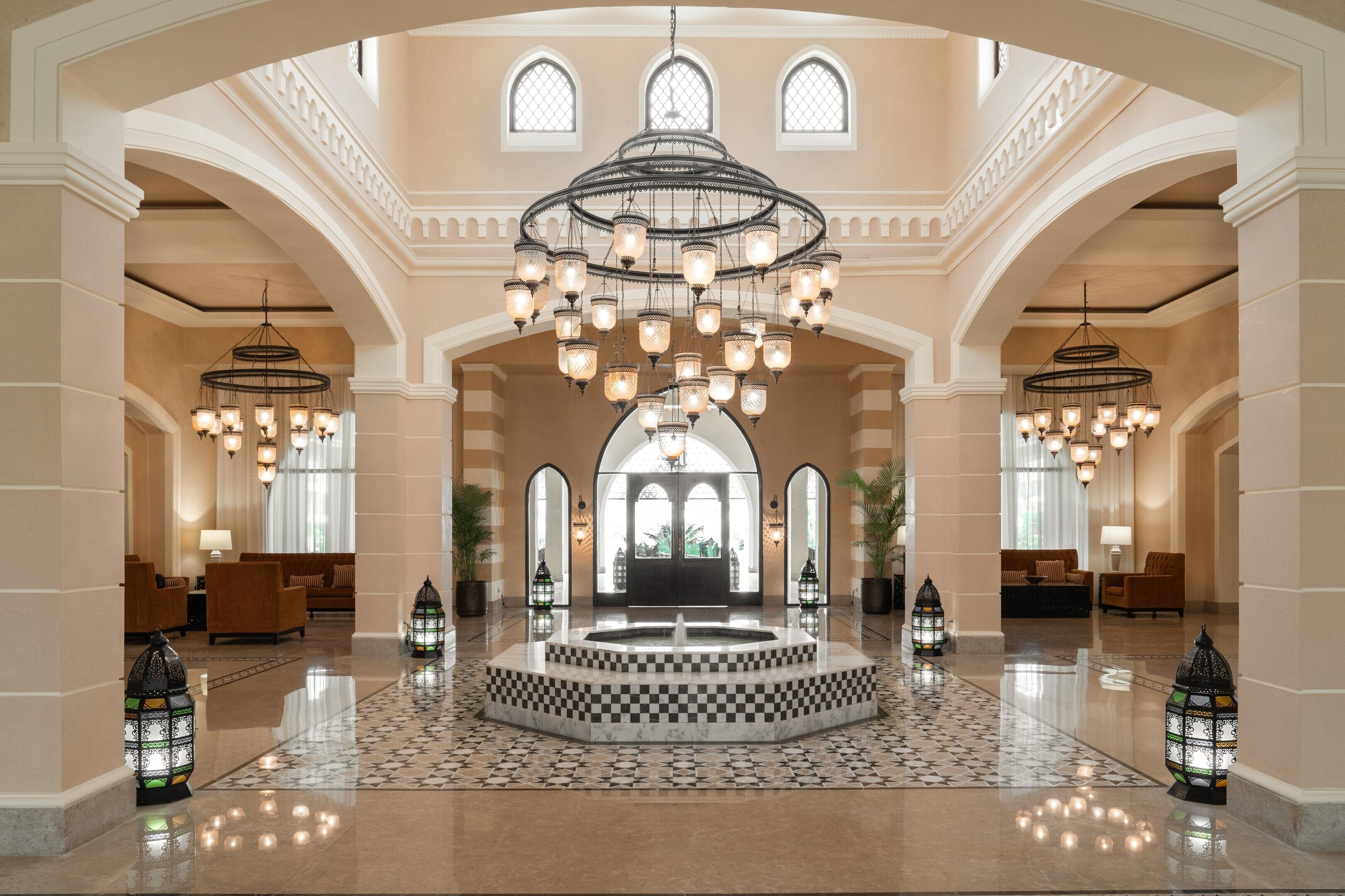 A spacious lobby with a central fountain, arched doorways, and multiple hanging chandeliers.
