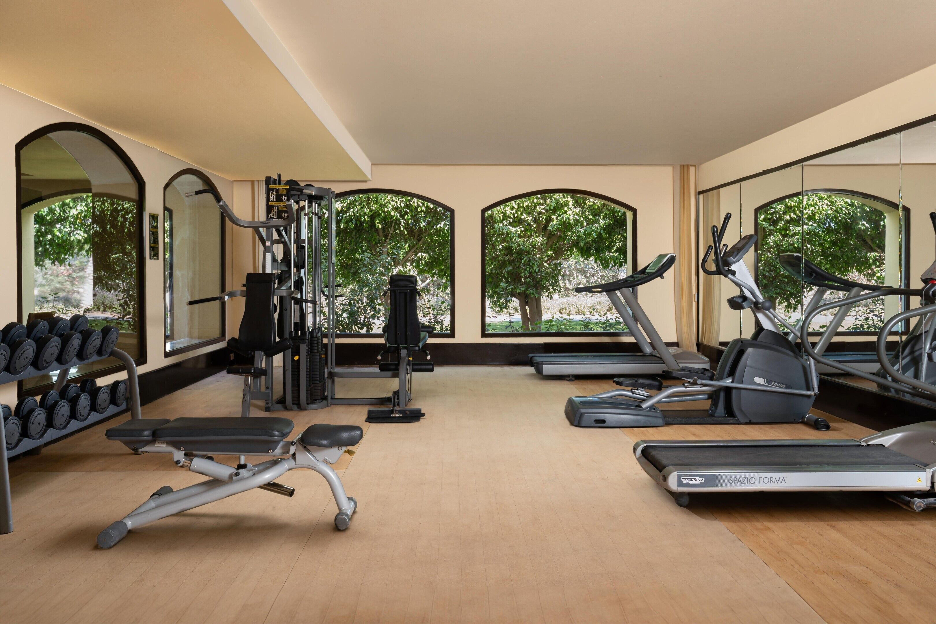 Un gimnasio con varios equipos de ejercicio, amplias ventanas con vista a árboles y suelo de madera.