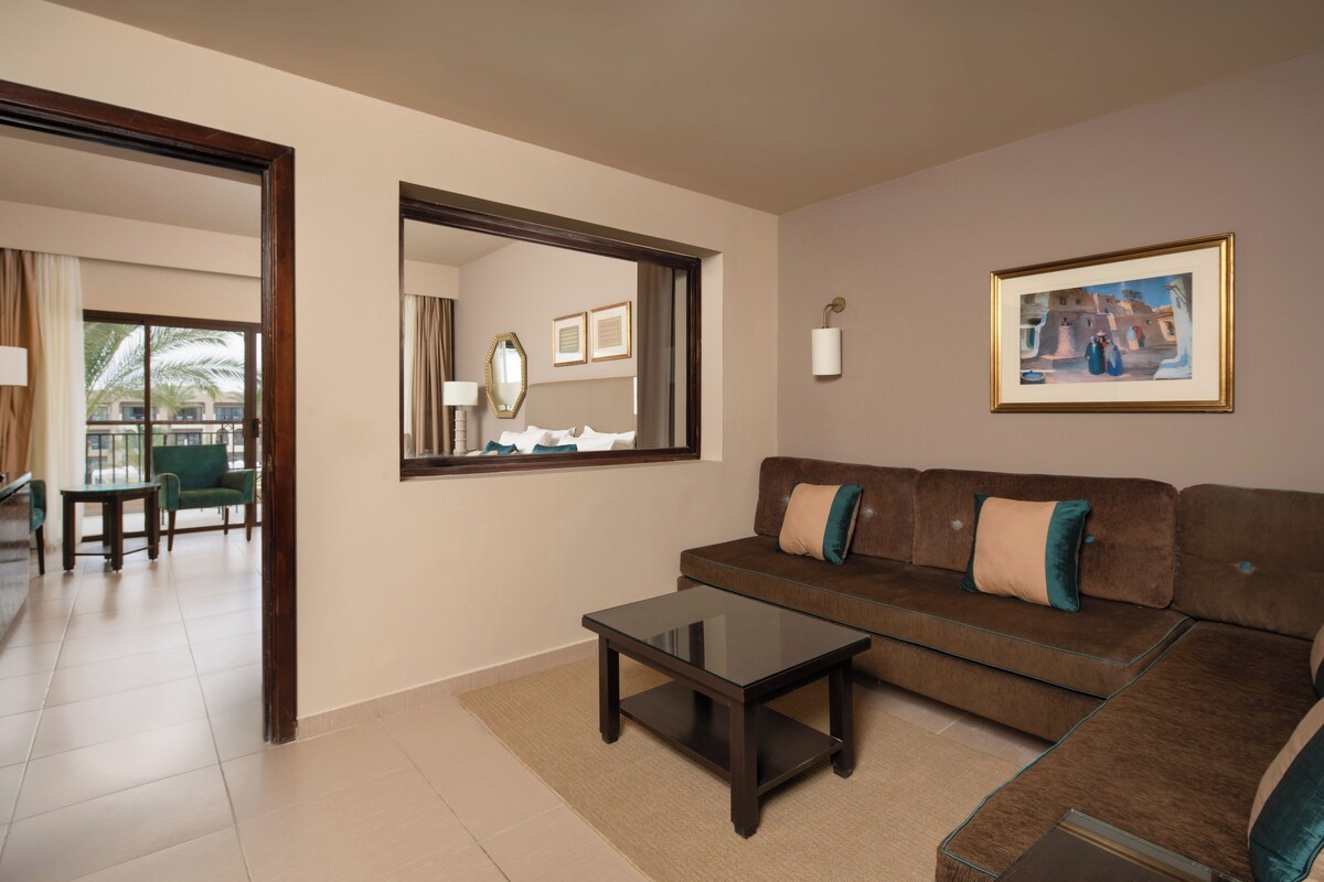 A modern living room with a brown sofa, a glass coffee table, and a large mirror. There is a framed picture on the wall and a view of the outside balcony.