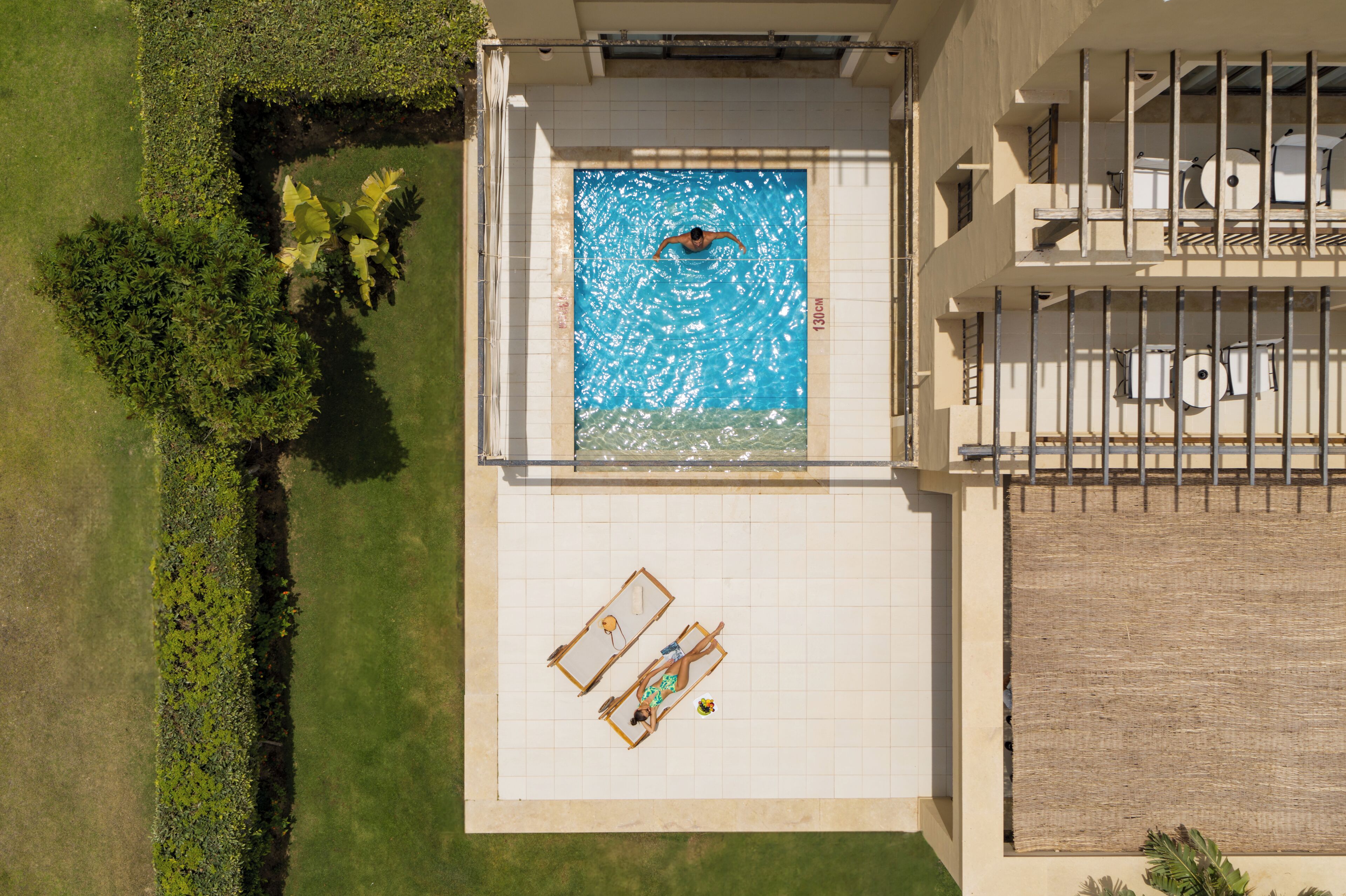 Eine Vogelperspektive auf einen Swimmingpool mit einer schwimmenden Person, umgeben von einer gefliesten Liegefläche und einer kleinen Lounge-Area.