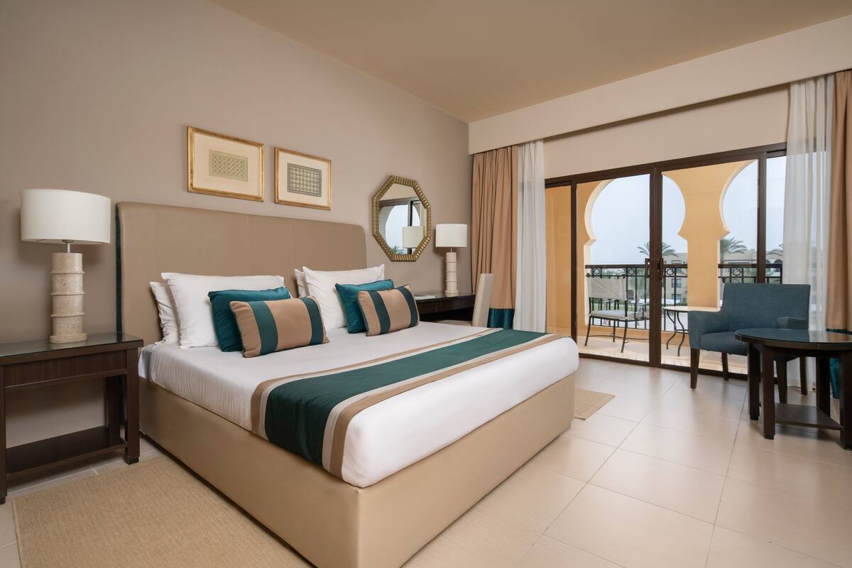 Ein Hotelzimmer mit einem großen Bett, zwei Nachttischen, einem Sessel und einem Balkon mit Blick, sowie einem großen Fenster.