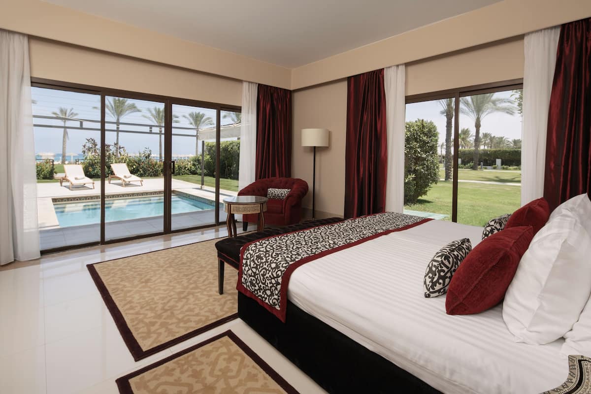 Ein geräumiges Schlafzimmer mit einem großen Bett, einem roten Sessel und Blick auf Pool und Garten.