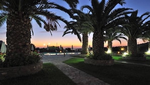 A pathway lined with palm trees and a stone walkway leading to a body of water at dusk. - Eri Hotel (Paros)