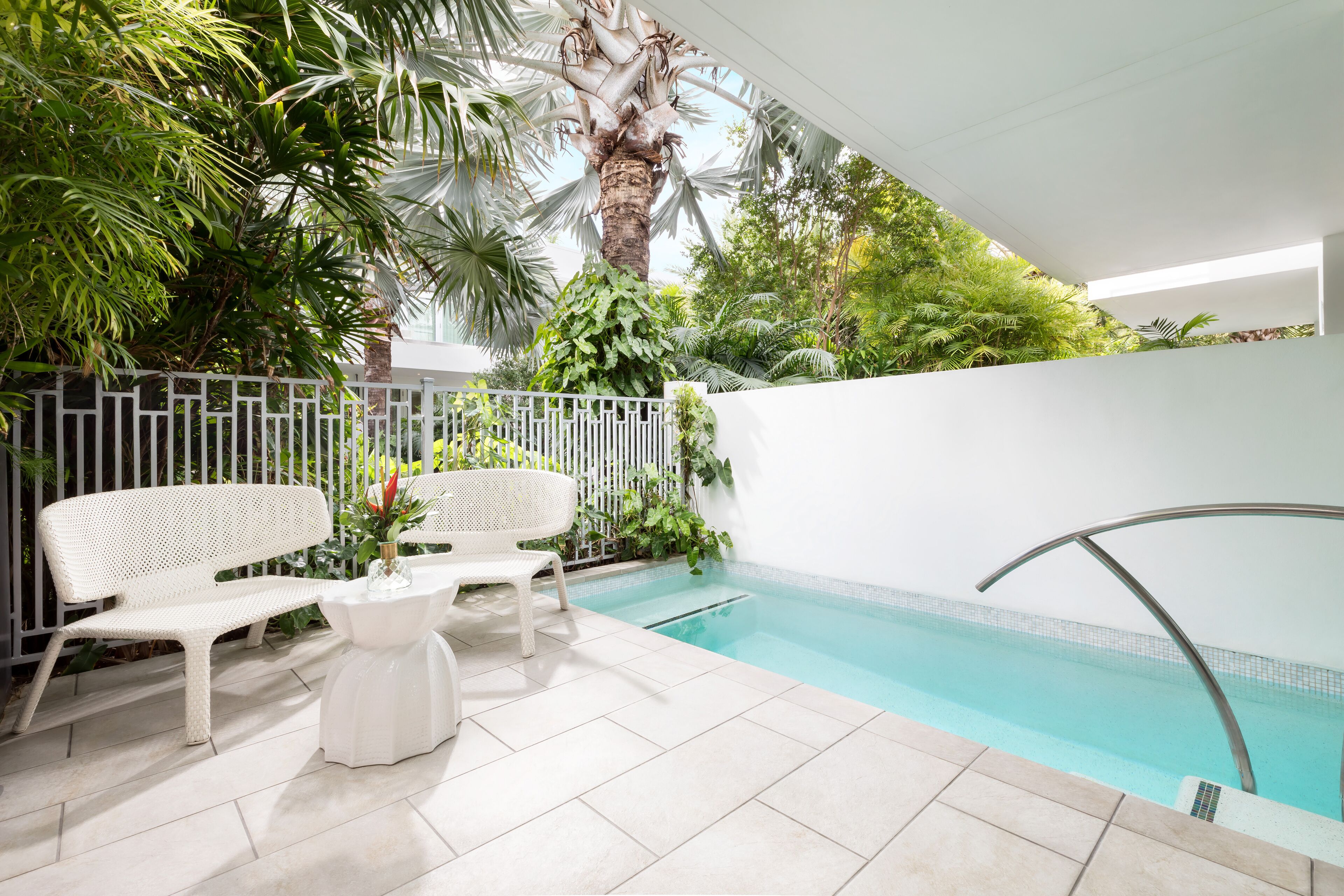 A pool area with two white wicker chairs and a small table, surrounded by lush greenery and a white fence.