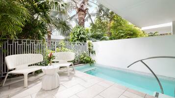 A pool area with two white wicker chairs and a small table, surrounded by lush greenery and a white fence.