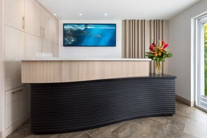 A modern reception desk with a wooden top and black base, a large TV screen above, and a vase with flowers to the right. - H2O Suites - Adults Only (Key West)