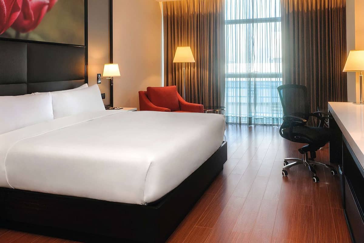 A hotel room with a large bed, two bedside lamps, a desk with a chair, and a large window with curtains.