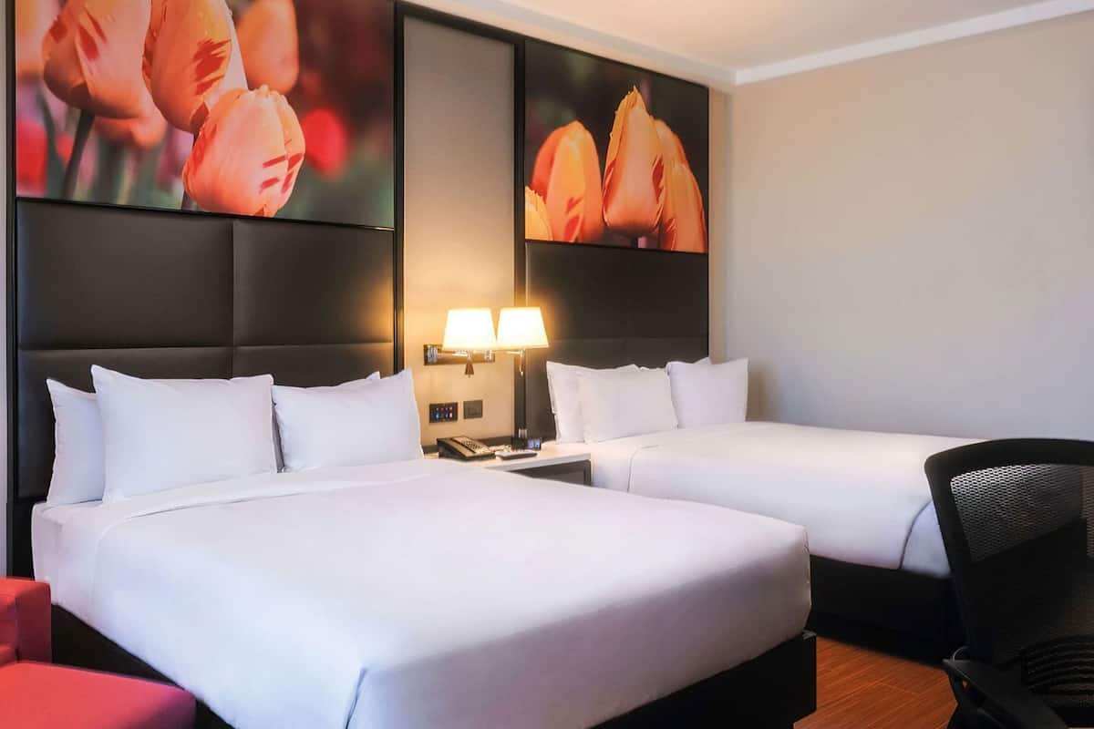 A hotel room with two beds, a red chair, a desk, and a wall-mounted lamp.