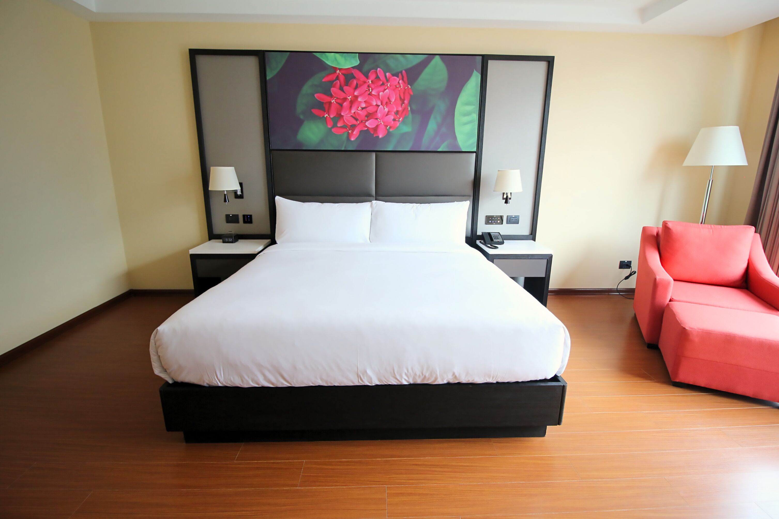 Une chambre d’hôtel avec un grand lit, deux tables de chevet avec des lampes, un fauteuil rouge et une grande œuvre d’art à motifs floraux accrochée au mur.