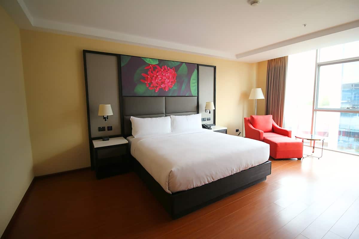 A modern hotel room with a large bed, a red armchair, a nightstand with a lamp, and a large window offering a view of the city.